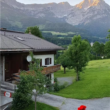 Ein malerisches Haus in den Bergen mit einer schönen Aussicht auf die umliegenden Gipfel. Der Garten ist üppig grün und mit roten Blumen geschmückt.