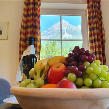 Eine Schüssel mit frischem Obst, darunter Äpfel, Trauben und Bananen, steht auf einem Tisch. Im Hintergrund sieht man ein Fenster mit einer atemberaubenden Berglandschaft.