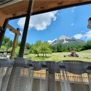 Ein schöner Blick aus dem Fenster auf die Berge und eine grüne Wiese. Im Vordergrund ist ein Traktor zu sehen, der in der Landschaft arbeitet.