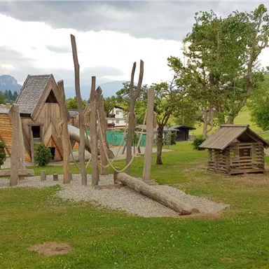 Ein Spielplatz mit Holzstrukturen, Schaukeln und einem kleinen Haus. Die Umgebung ist grün mit Bäumen und Bergen im Hintergrund.