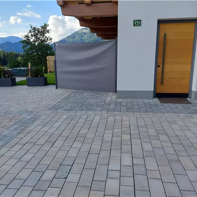 Ein gepflasterter Eingangsbereich mit einer Holztür und einem Sichtschutz. Im Hintergrund sind Bäume und Berge zu sehen.