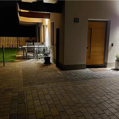 Ein moderner Eingangsbereich mit einer gepflasterten Fläche. Im Hintergrund sind Gartenmöbel und eine Rasenfläche sichtbar.