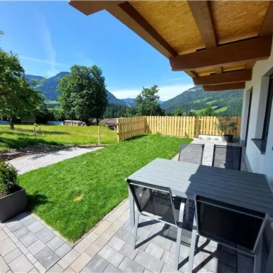 Ein schöner Außenbereich mit einer Terrasse, einem Tisch und Stühlen. Im Hintergrund sind grüne Wiesen und Berge zu sehen.