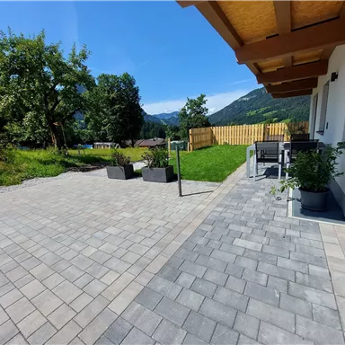 Ein schöner Außenbereich mit gepflastertem Boden und modernen Möbeln. Im Hintergrund sind Bäume und Berge sichtbar, unter einem klaren blauen Himmel.