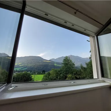 Ein offenes Fenster mit Blick auf grüne Wiesen und Berge. Der Himmel ist klar und die Landschaft ist ruhig und idyllisch.