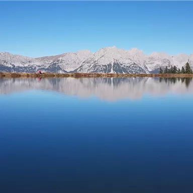 Ein ruhiger, klarer See spiegelt die Berge und den blauen Himmel wider. Die Landschaft ist von Bäumen umgeben und vermittelt eine friedliche Atmosphäre.