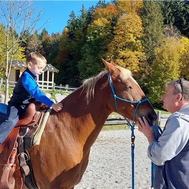 Ein Junge sitzt auf einem Pferd und spricht mit einem Erwachsenen. Im Hintergrund sind Bäume mit buntem Laub und eine Reitstation zu sehen.