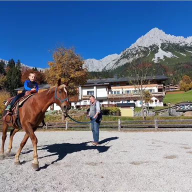 Ein Kind sitzt auf einem Pferd, während eine Person es an der Leine hält. Im Hintergrund sind Berge und ein Bauernhaus zu sehen.