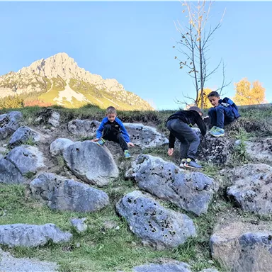 Drei Kinder spielen und klettern an einem felsigen Hanggelände. Im Hintergrund sind majestätische Berge und ein klarer Himmel zu sehen.