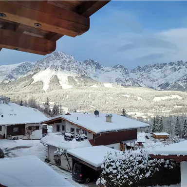 Eine verschneite Landschaft mit Bergkulisse und traditionellen Chalets. Der Himmel ist blau und es liegt eine ruhige winterliche Atmosphäre in der Luft.