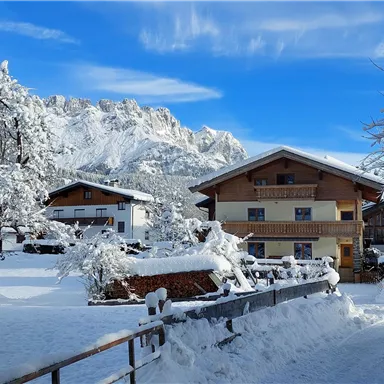 Ein malerisches Winterdorf mit verschneiten Häusern und schneebedeckten Bäumen. Im Hintergrund sind beeindruckende Berge und ein blauer Himmel zu sehen.