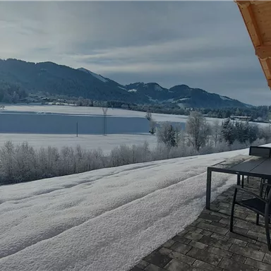 A snow-covered landscape with mountains in the background. On the terrace, there is a table and chairs, perfect for relaxing.