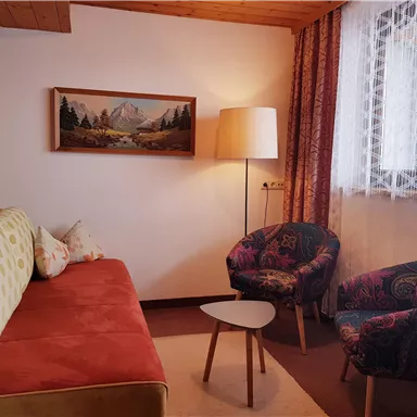 Ein gemütlicher Wohnraum mit einem roten Sofa und zwei bunten Sesseln. Ein Tisch und ein großes Fenster sorgen für viel Licht.