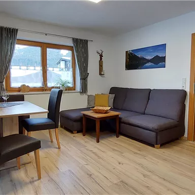 Ein gemütlicher Wohnraum mit einem Essbereich und einer Sofa-Ecke. Große Fenster bringen viel Tageslicht und bieten einen Blick auf die Umgebung.