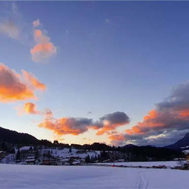 Eine verschneite Landschaft mit Bergen und Häusern im Vordergrund. Der Himmel ist von orangefarbenen Wolken durchzogen.