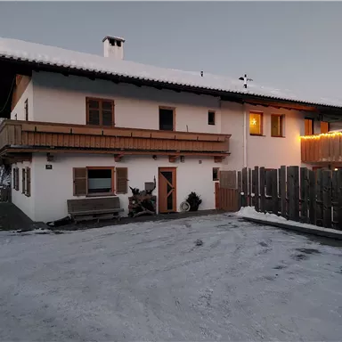 A cozy house in the snow with a balcony and warm light. The parking lot is covered in snow and there is a wooden fence.
