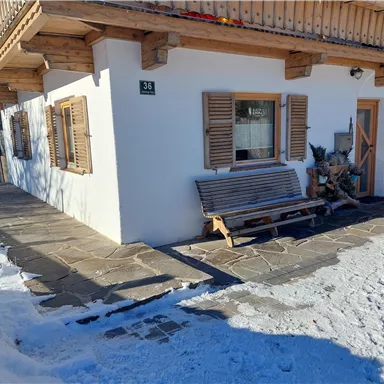 A cozy house in the snow with wooden shutters and a bench in front of the door. The path is paved with stones and the surroundings are wintry.