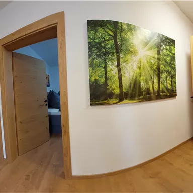 A bright hallway with wooden floors and open doors to several rooms. On the wall hangs a picture of a sunlit forest.