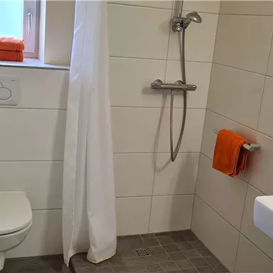Ein modernes Badezimmer mit einer Dusche und einem weißen Waschbecken. Handtücher in Orange und eine helle, minimalistische Gestaltung sorgen für eine freundliche Atmosphäre.