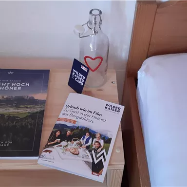 Ein Nachttisch mit einem Buch, einer Flasche Wasser und einer Broschüre. Der Tisch steht neben einem Bett mit kariertem Kissen.