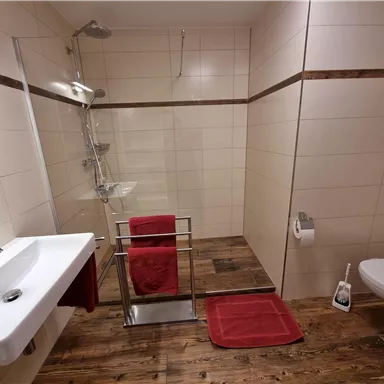 Ein modernes Badezimmer mit Dusche und Waschbecken. Rote Handtücher und eine Holzoptik am Boden sorgen für eine gemütliche Atmosphäre.