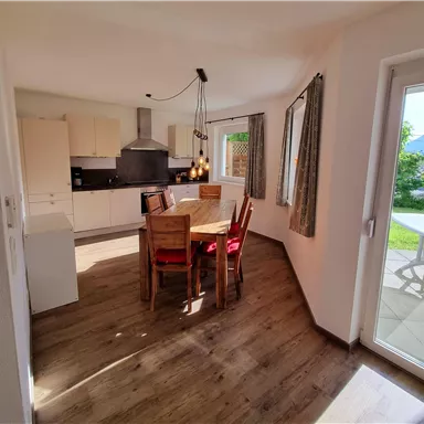 Eine helle Küche mit einem Essbereich und Holzfußboden. Der Tisch ist mit Stühlen ausgestattet, und es gibt große Fenster mit Blick nach draußen.