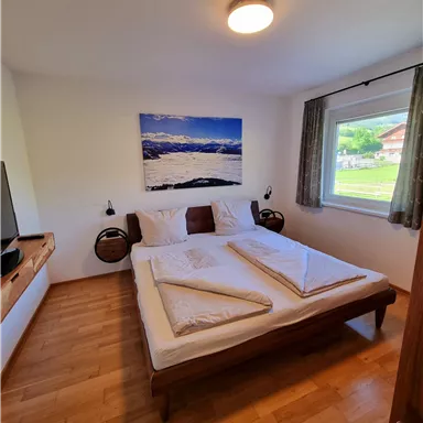 Ein helles Schlafzimmer mit einem Doppelbett und Holzfußboden. An der Wand hängt ein Bild von einer Landschaft, und es gibt ein Fenster mit Blick ins Freie.