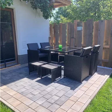 A cozy outdoor area with a table and chairs made of rattan. The area is surrounded by a wooden wall and has green grass.