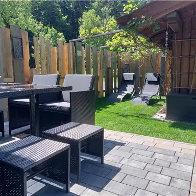 A cozy outdoor area with a table and chairs made of black wicker. In the background, deck chairs and a small wooden shed are visible, surrounded by green grass.