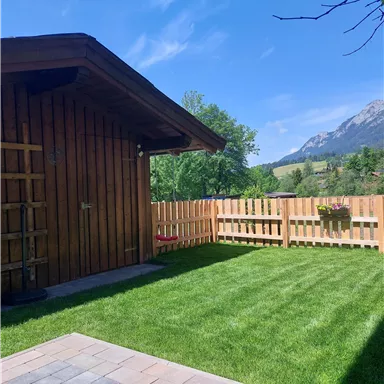 A cozy wooden house with a well-maintained garden. In the background, green trees and mountains can be seen under a blue sky.