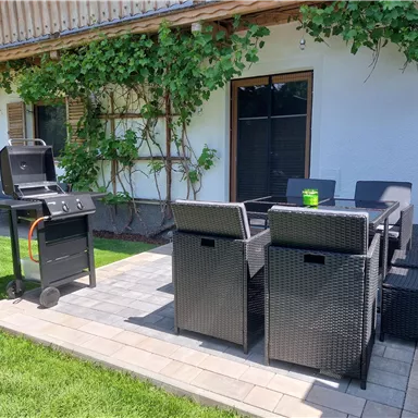 A cozy terrace with a grill and modern garden chairs. In the background, green plants and a bright house wall can be seen.