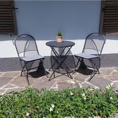 A small outdoor area with a round table and two folding chairs. A small plant stands in the center of the table, surrounded by green grass.