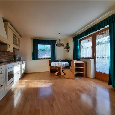 Eine helle und gemütliche Küche mit Holzfußboden. Es gibt einen Tisch, Stühle und große Fenster mit grünen Vorhängen.