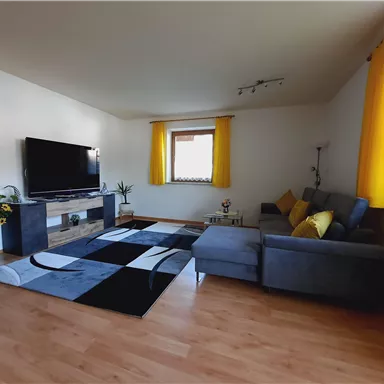 Ein modernes Wohnzimmer mit einem großen Fernseher und einem gemütlichen Sofa. Gelbe Vorhänge und ein stilvoller Teppich sorgen für eine einladende Atmosphäre.