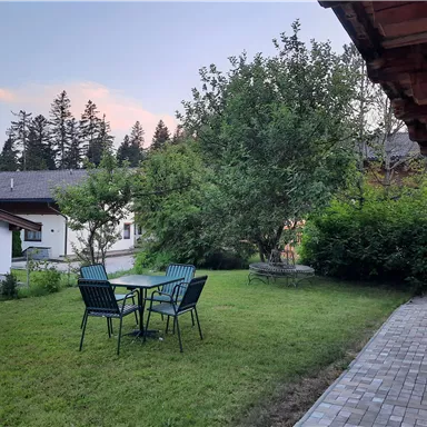 Ein ruhiger Garten mit einem Tisch und Stühlen, umgeben von Bäumen und Sträuchern. Im Hintergrund sind einige Gebäude sichtbar.