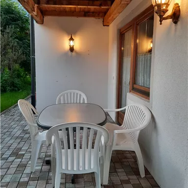 Ein gemütlicher Außenbereich mit einem Tisch und vier Kunststoffstühlen. Die sanfte Beleuchtung sorgt für eine entspannte Atmosphäre.