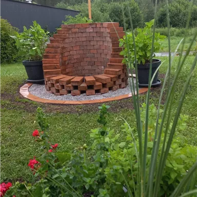 Ein dekorativer gemauerter Sitzplatz in einem Garten, umgeben von Grünpflanzen und Blumen. Im Hintergrund sind Berge und Bäume sichtbar.