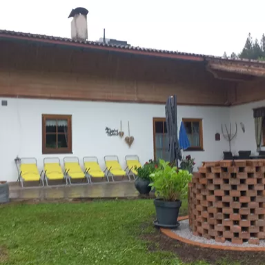 Ein traditionelles Haus mit einem Holzverand und einem Garten. Vor dem Haus stehen gelbe Liegestühle und Pflanzen in Töpfen.