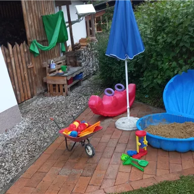 Ein Spielbereich im Freien mit einer Sandkiste, Schaufeln und einer bunten Rutsche. Hintergründe sind grün und ein Sonnenschirm steht bereit.