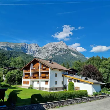 Ein gemütliches Haus in den Bergen, umgeben von grüner Wiese und Bäumen. Im Hintergrund sind hohe Berge unter einem klaren blauen Himmel zu sehen.