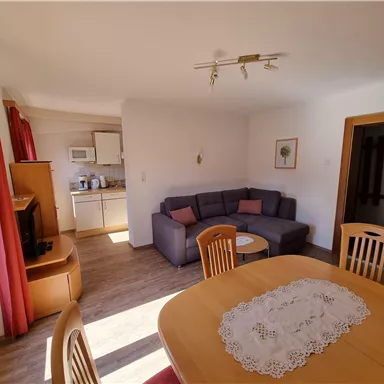 Ein gemütliches Wohnzimmer mit einer Couch und einem Esstisch. Die Küche ist im Hintergrund sichtbar und große Fenster lassen viel Licht hereinscheinen.