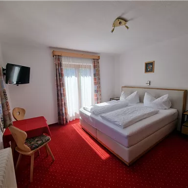 Ein gemütliches Schlafzimmer mit einem Doppelbett und zwei weißen Kopfkissen. Es gibt einen Tisch, einen Stuhl und einen Fernseher, sowie viel Tageslicht von den Fenstern.