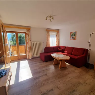 Ein gemütliches Wohnzimmer mit einem roten Sofa und einem runden Tisch. Helle Fenster bieten einen Blick nach draußen und viel Tageslicht.