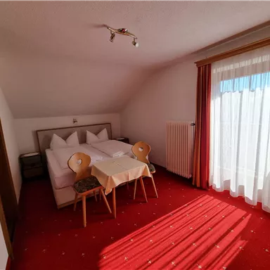 Ein gemütliches Zimmer mit einem Doppelbett und einem kleinen Tisch. Viel Tageslicht fällt durch die Fenster auf den roten Teppich.