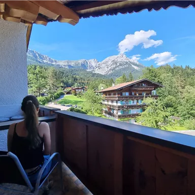 Eine Person sitzt auf einem Balkon und blickt auf eine malerische Berglandschaft. Im Vordergrund stehen Bäume und ein Gebäude, das die Natur umrahmt.