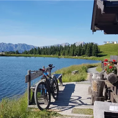 Ein gemütliches Holzhäuschen am Ufer eines ruhigen Sees. Im Vordergrund stehen Fahrräder und im Hintergrund sind Berge und Wiesen zu sehen.