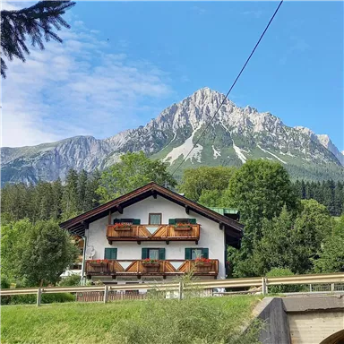 Ein charmantes Haus in den Alpen mit Balkonen und grünem Umfeld. Im Hintergrund erhebt sich ein majestätischer Berg unter einem blauen Himmel.