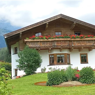 Ein schönes, traditionelles Haus mit einem Holzbalon und bunten Blumen im Garten. Im Hintergrund sind Berge und eine grüne Landschaft zu sehen.
