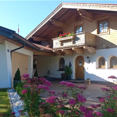 Ein schönes, traditionelles Haus mit Holzverkleidung und einem Balkon. Vor dem Haus blühen bunte Blumen in einem gepflegten Garten.