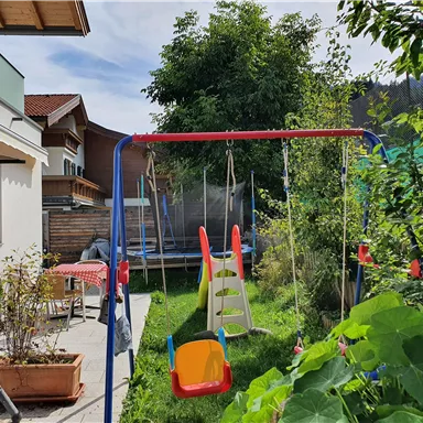 Ein schöner Garten mit einer Schaukel und einer Rutsche. Im Hintergrund ist ein Trampolin und einige grüne Pflanzen zu sehen.